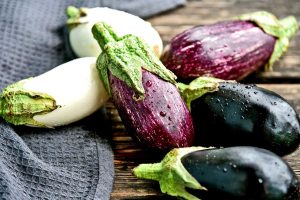 Three different types of eggplant in a pile on rustic table; purple, dark purple, and white colored fruit.