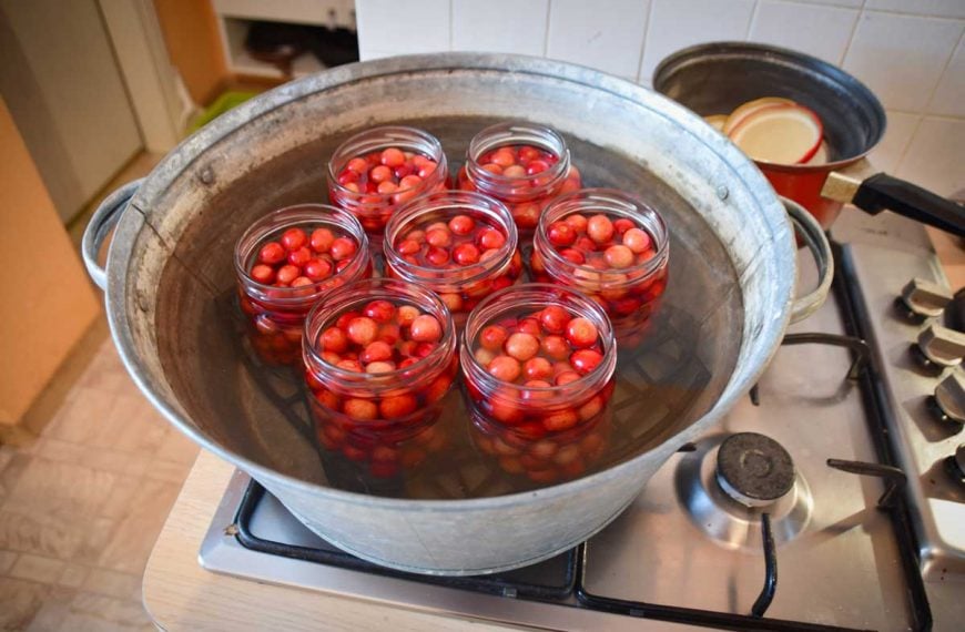 Cherry compote being prepared for preservation by using a double boiler method with canning jars inside of a galvanized tub on a stovetop.